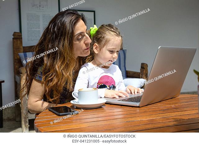 girl four years old sitting on mother legs, surfing internet in notebook computer together, with coffee cup and black mobile smart phone on brown wooden table