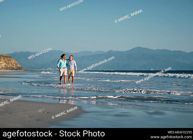 Mature gay men holding hands while walking on shore at beach against clear blue sky, Riviera Nayarit, Mexico