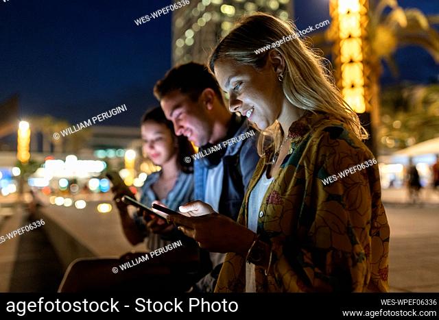 Smiling woman surfing net through mobile phone sitting by friends at night