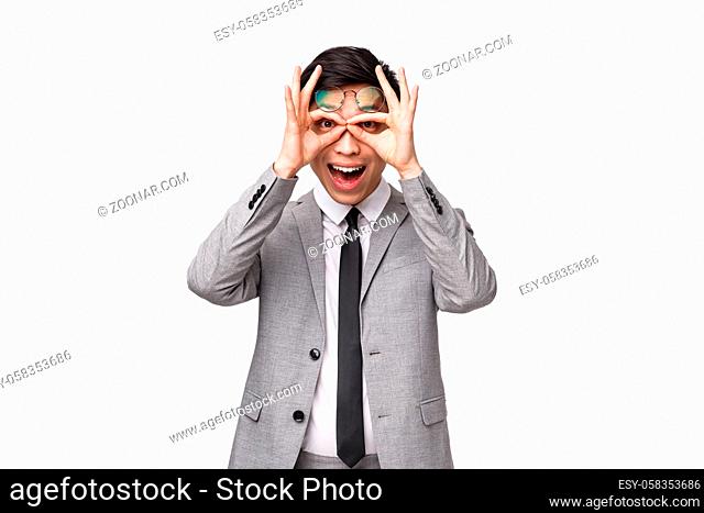 Waist-up portrait of funny and amused, excited asian male entrepreneur in business suit, making glasses with fingers and looking through it at camera with...