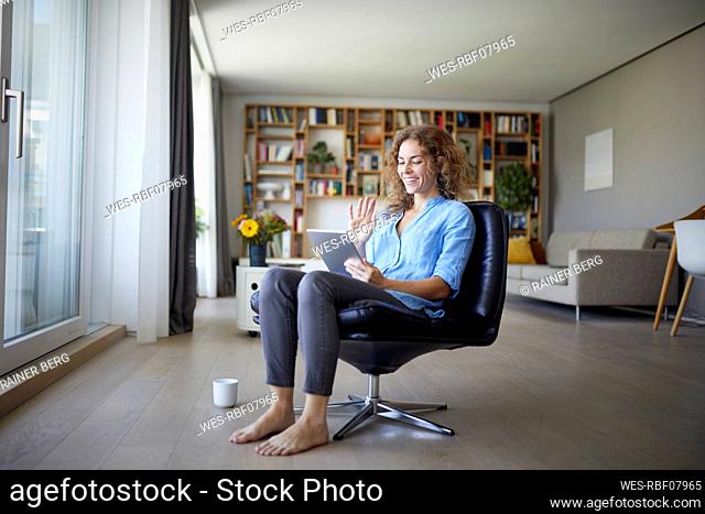 Smiling woman waving hand to video call on digital tablet while sitting on chair at home