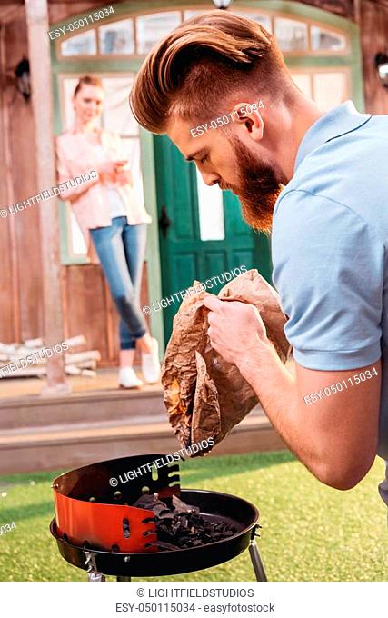Young woman standing on porch and looking at bearded man holding coal for outdoor grill
