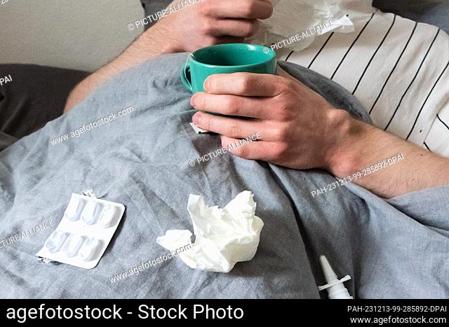 12 December 2023, Lower Saxony, Hanover: ILLUSTRATION - A man lies in bed with handkerchiefs, teacup, nasal spray and tablets (staged scene)