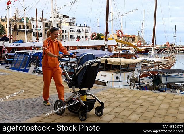CYPRUS, KYRENIA - DECEMBER 15, 2023: A woman pushes a chile in a pram on a promenade by a marina in winter. The Turkish Republic of Northern Cyprus is a de...
