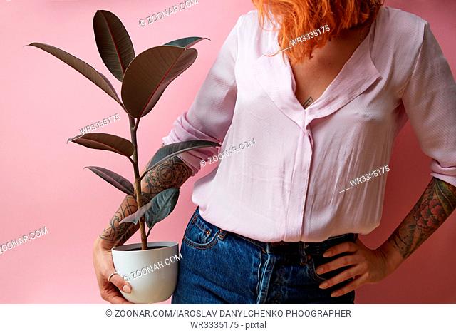 Red-haired girl with a tattoo holding a vase with a ficus on a pink background. Flowers shop concept