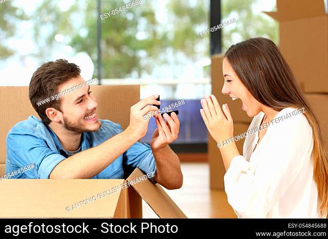 Happy man proposing to excited girlfriend with wedding ring while moving at new home surrounded by boxes