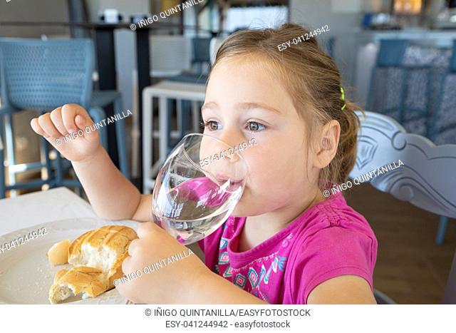 four years old blonde little girl with pink shirt drinking water from cup, sitting indoor in restaurant