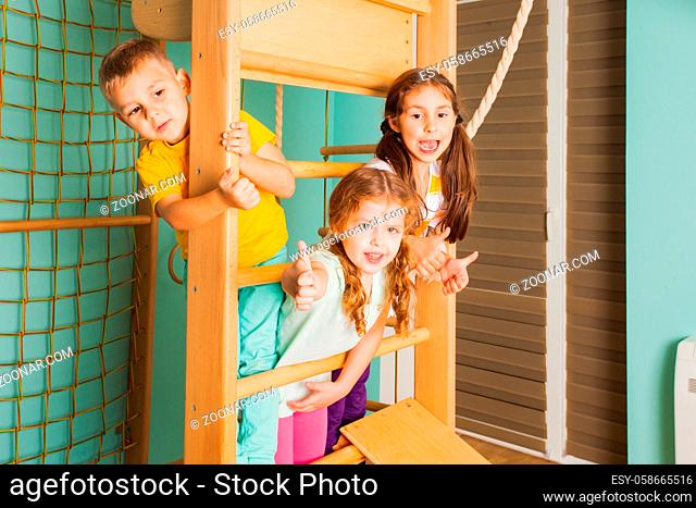 Portrait of cheerful smiling children on a wooden swedish wall, holding on, looking at the camera. Two adorable girls and cute boy smiling