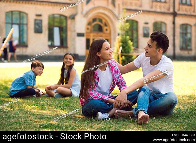 Young Teachers Sitting On Grass In School Yard. Two Kids Sitting Behind Them. Relaxing Time Before Summer Holidays