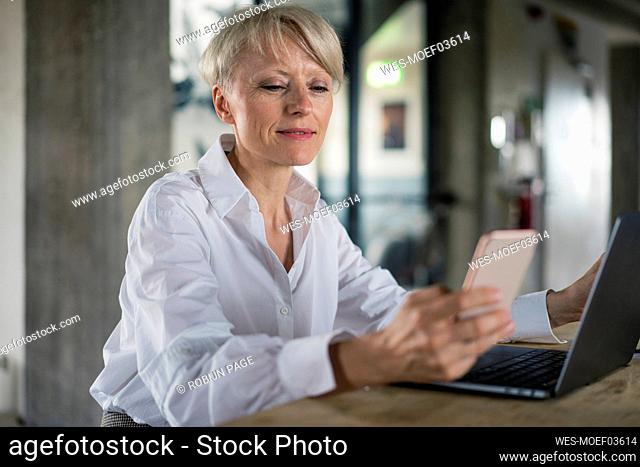 Blond businesswoman with laptop using mobile phone while sitting at desk in home office