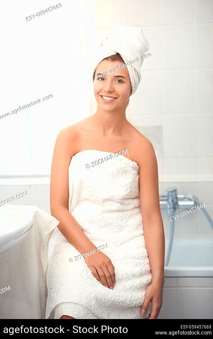 Pretty healthy young girl wrapped in white towels standing in a bathroom with a happy smile in a spa treatment, wellness and personal hygiene concept