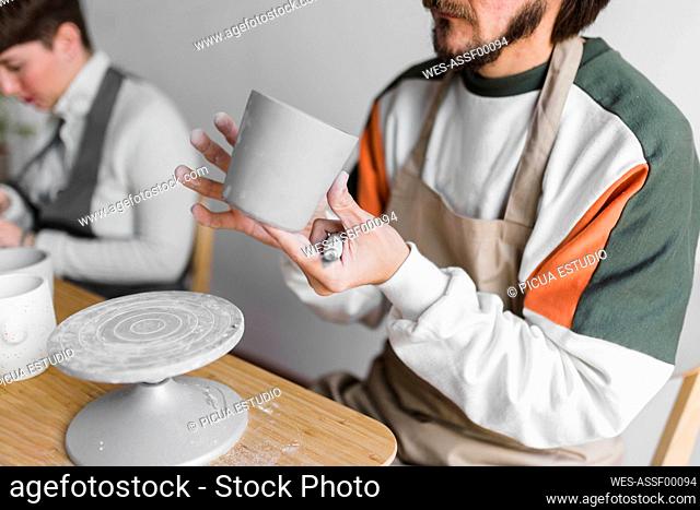 Male craftsperson checking clay cup while working with colleague at workshop