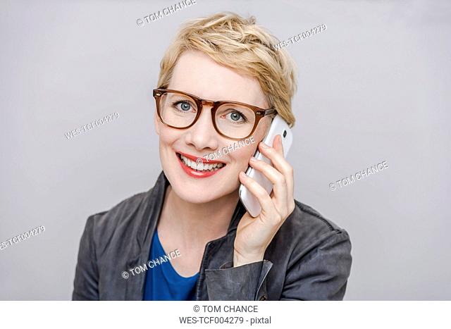 Portrait of smiling blond woman wearing glasses telephoning with smartphone in front of grey background