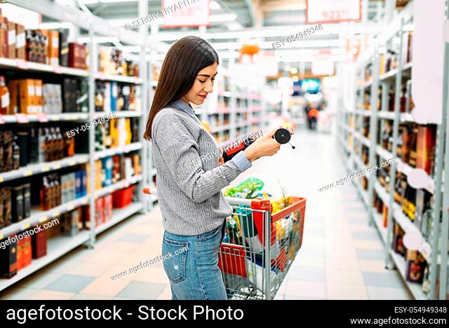 Woman with cart in supermarket, alcohol drinks department, family shopping. Female customer choosing alcoholic beverages in shop, buyers in market