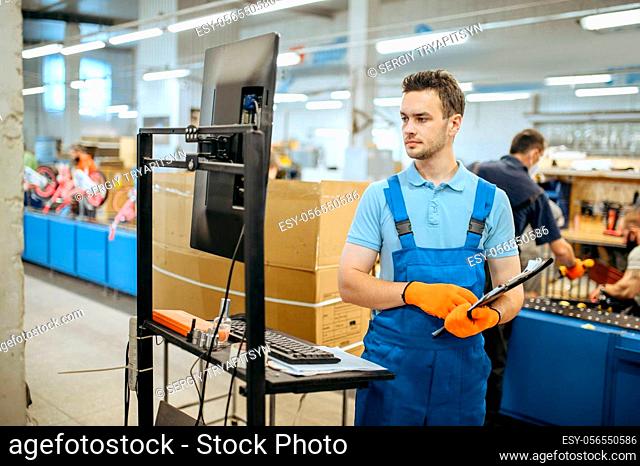 Bicycle factory, worker with notebook poses at bike assembly line. Male mechanic in uniform installs cycle parts in workshop, industrial manufacturing