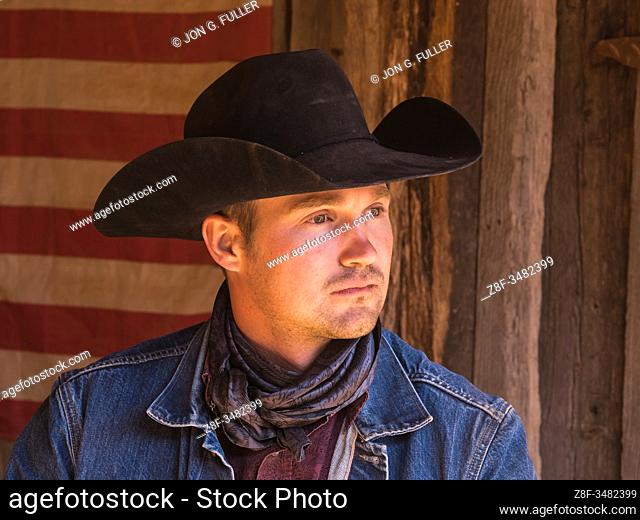 A working cowboy wrangler poses in his cowboy hat and bandana in front of an American flag on the Red Cliffs Ranch near Moab, Utah