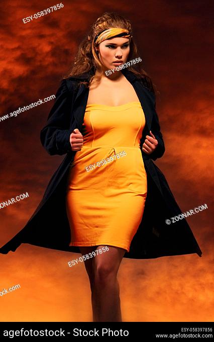Elegant beautiful woman wearing yellow dress and black coat tied a scarf on head posing against cloudy orange red cloudscape background