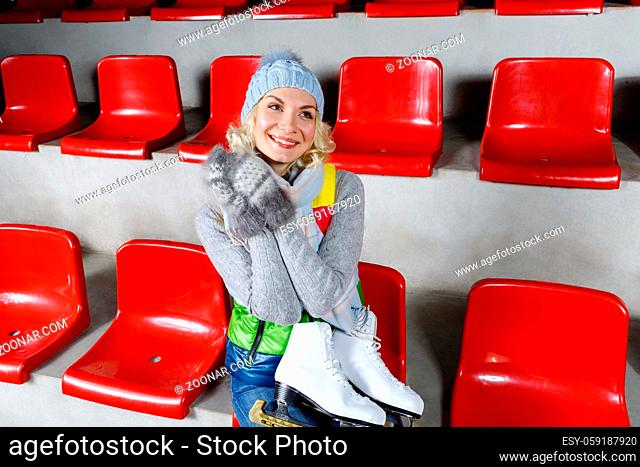 Beautiful blond young woman in winter clothes sitting on red tribune and holding white skates