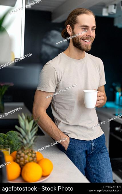 Smiling young adult man with beard holding cup with coffee, standing by kitchen counter with modern light interior, enjoying fresh warm drink at home