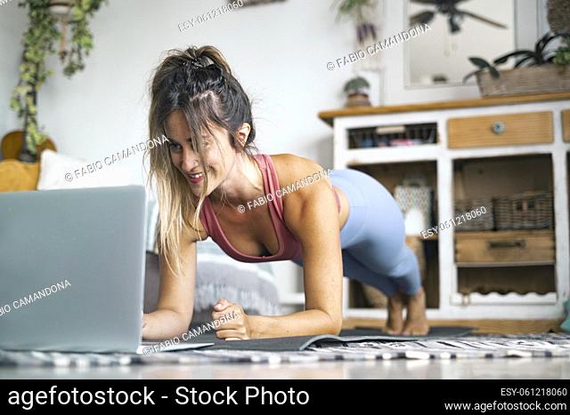 Beautiful young woman doing plank exercise while looking at laptop on floor. Woman in sportswear doing pilates at home. Woman in sportswear exercising at home...