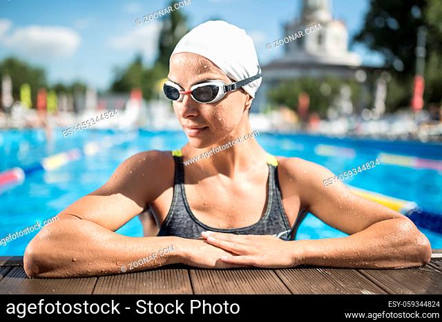 Happy girl in the multi-colored swimwear and swim glasses looks to the right in the swimming pool outdoors. She leans on the side of the pool with her hands and...