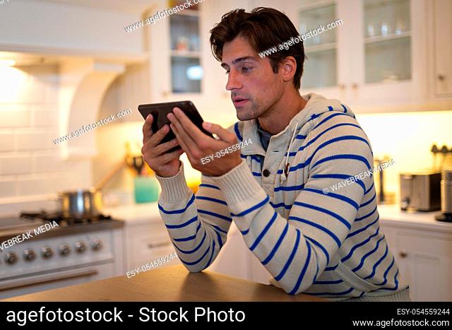 Side view of a Caucasian man at home wearing a blue striped sweater, sitting in the kitchen at the kitchen island, using a tablet computer and concentrating