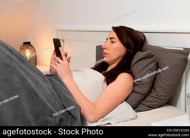 Beautiful woman lying in bed and checking mobile phone before going to sleep. Side view