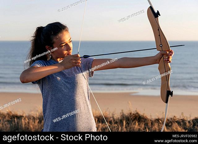 Young woman aiming with bow and arrow while standing at beach