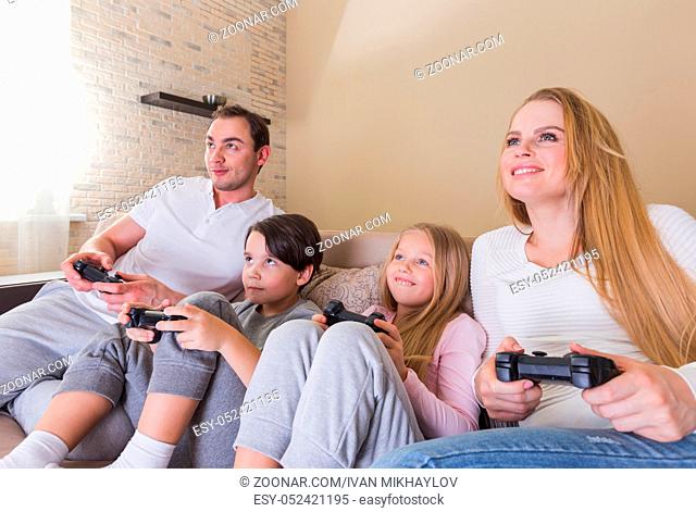 Family of parents and two children playing video game together sitting on sofa at home