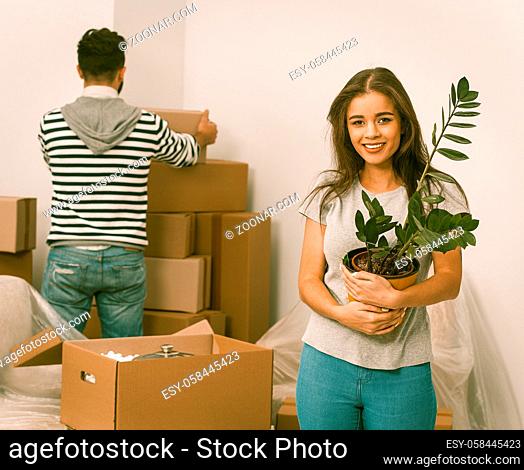 Young couple relocating to new apartment and unpacking boxes. Front view of young woman holding plant and man standing behind unpacking boxes