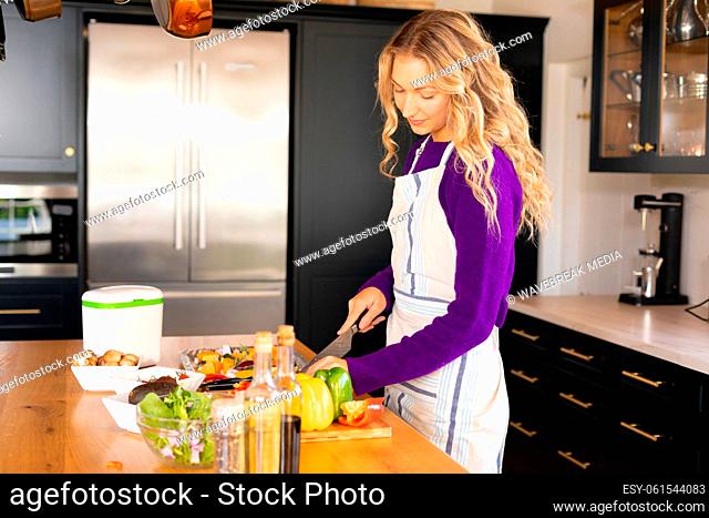 Happy caucasian woman standing in kitchen wearing apron chopping vegetables