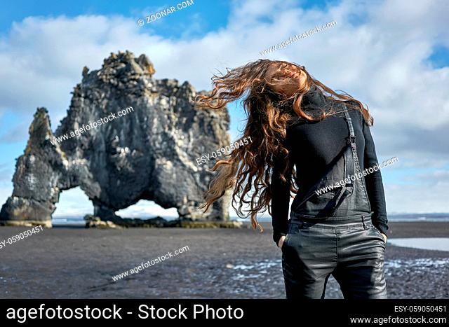 Pretty girl with windy hair stands on the volcanic beach on the background of the Hvitserkur in Iceland. She wears black jumper with dark overalls and holds...