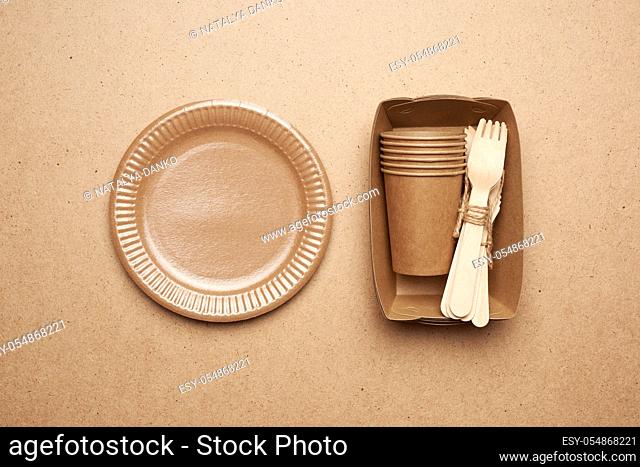 paper plates and cups from brown craft paper and wooden forks and knive on a brown background. Plastic rejection concept, zero waste