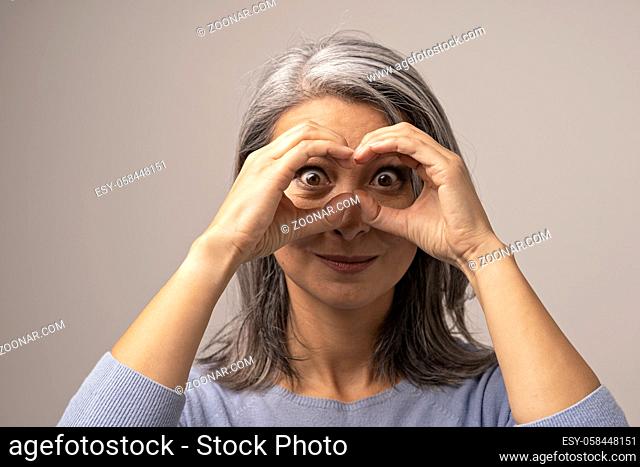 Mongolian Mature Woman Holds Hands As if Looking Through Binoculars. The Head of the Woman is Covered with Gray Hair. Her Eyes are Big and Brown