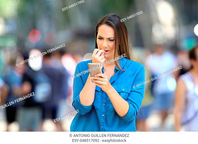 Nervous woman biting nails checking smart phone content standing in the street
