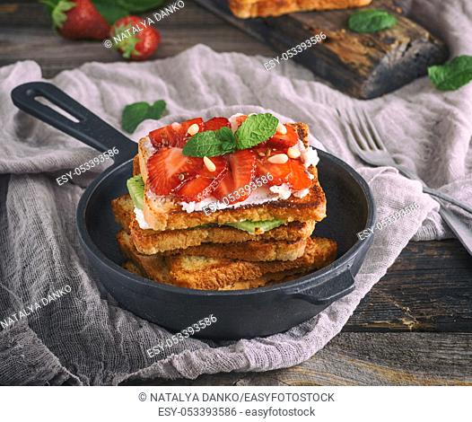 pile of French toast from white bread with cottage cheese, strawberries in a black cast-iron round frying pan with a handle on a gray wooden table