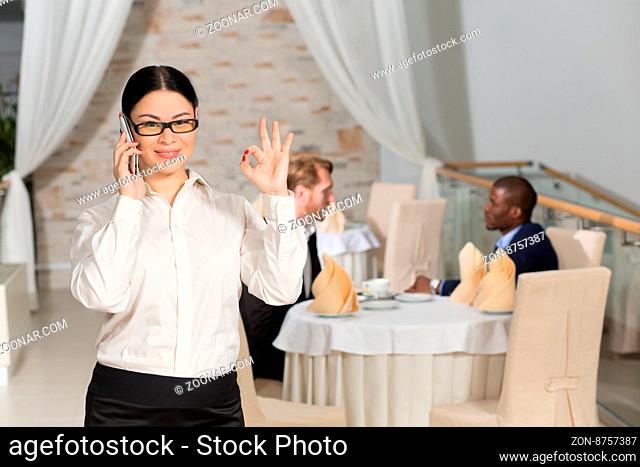 Happy business woman speaking over mobile phone and showing okay while having business meeting in restaurant