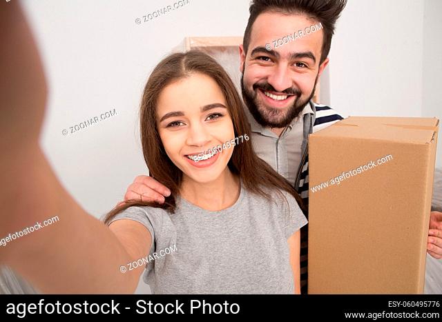 Close up portrait of young couple after relocation making selfie. Smiling man and woman making selfie shot, man is holding cardboard box