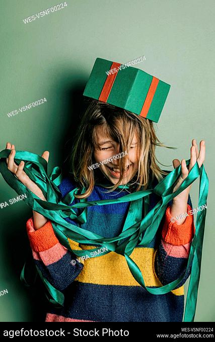 Girl playing with tangled Christmas ribbons and gift box in front of green wall