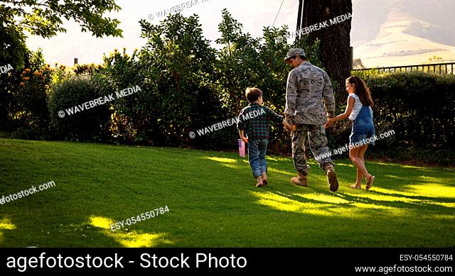 Rear view of a young adult mixed race male soldier in the garden outside his home, holding hands and walking with his young son and daughter