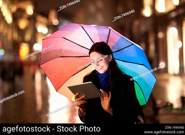 Happy young girl with colorful umbrella using tablet computer outdoor. She standing in the city street in the rainy evening