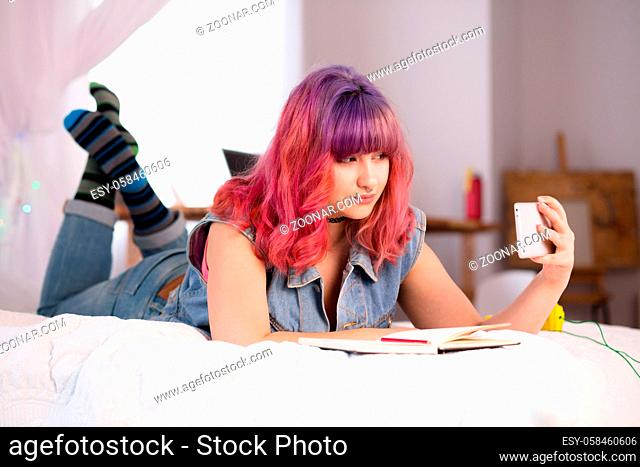 Hipster girl lying on bed and making self photos on mobile or smart phone at home. Lady with pink hair posing for camera. Hipster concept
