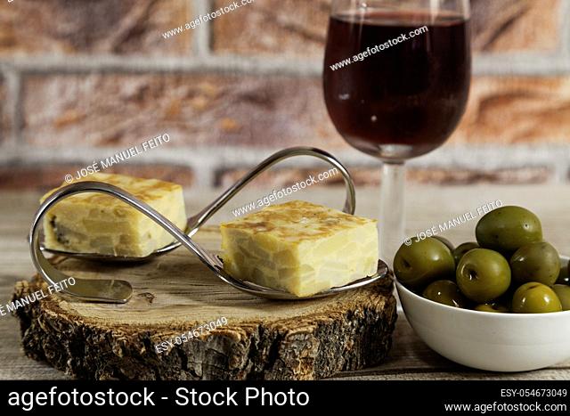 Spanish omelette brochettes on a wood slice, olives bowl and red wine glass on wooden table and red brick background