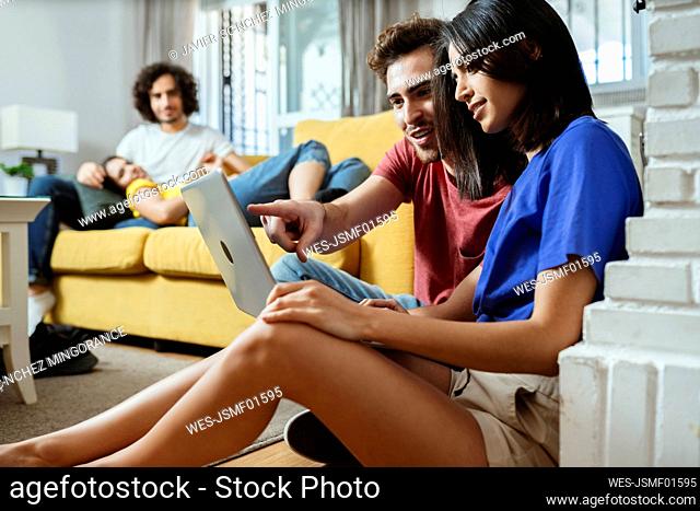 Young couple sharing laptop while friends relaxing on sofa in living room at home