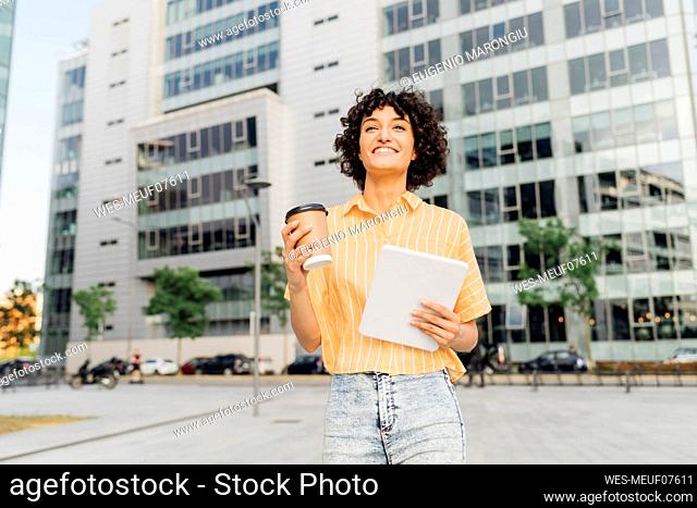 Smiling businesswoman with reusable coffee cup and tablet PC walking in front of building