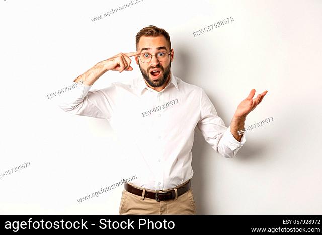 Confused and shocked man pointing at head, scolding employee for acting stupid, standing over white background
