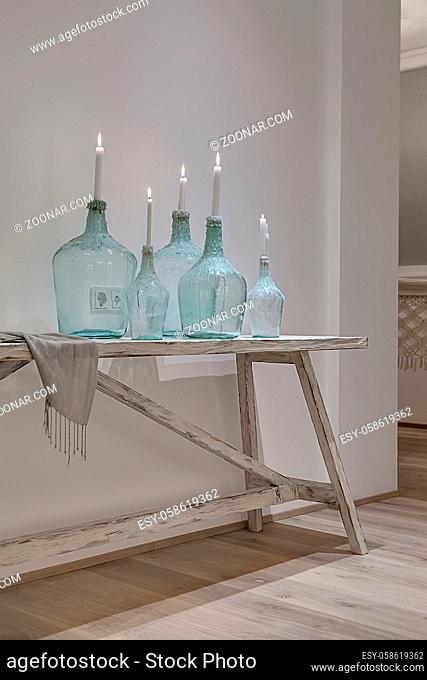 Glowing candles in the stylish bottles on the wooden table on the white wall background. There is a shawl near them. Behind the bottles there are power sockets...