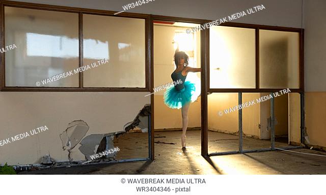 Front view of a young mixed race female ballet dancer wearing a blue tutu and pointe shoes dancing on on leg in a doorway at an abandoned warehouse building