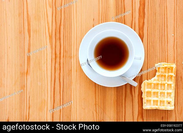 Wooden table with white cup with saucer and spoon as well as lying waffle with bite mark in top view