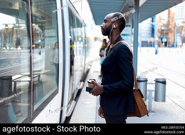 Young businessman with coffee to go standing at tram stop listening music with earphones and smartphone